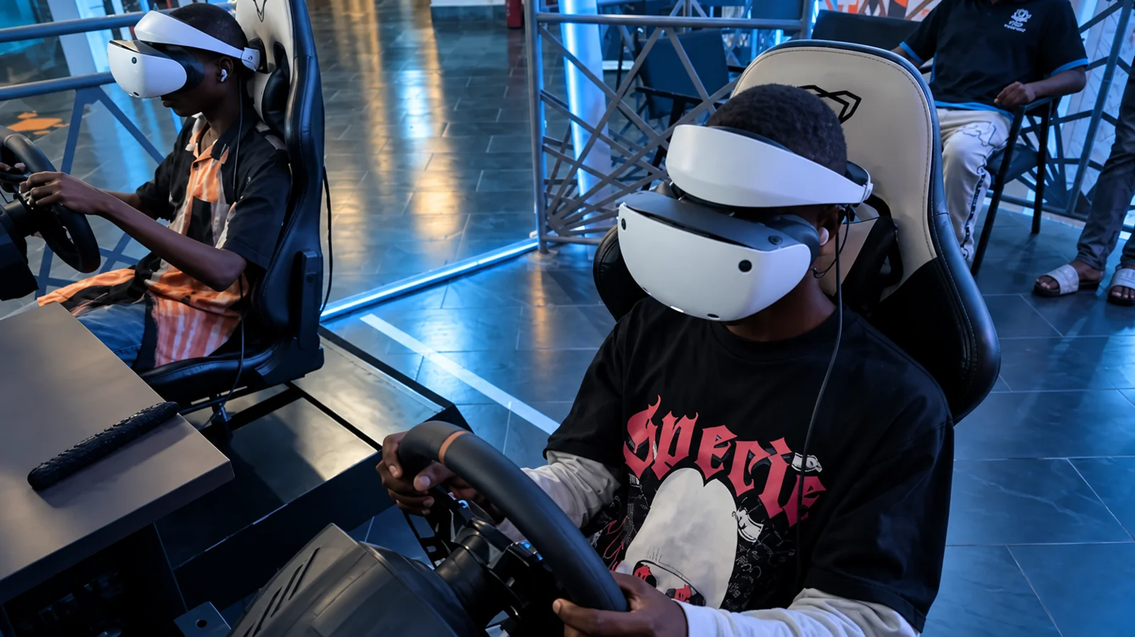 Two Player One racers wearing VR headsets at their racing rigs, with edited lighting